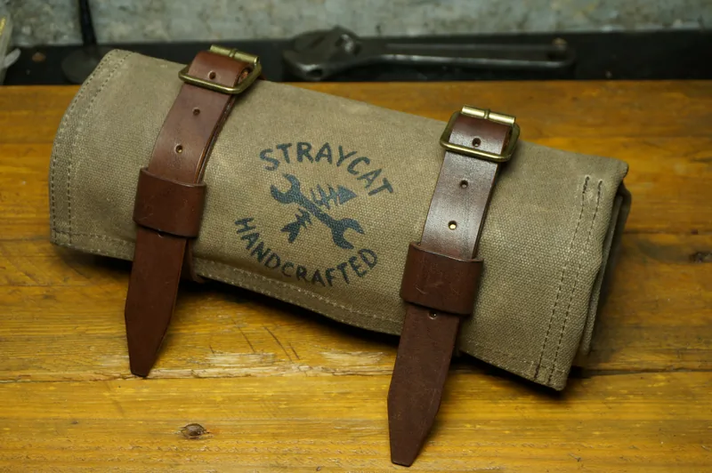 Tool Roll „Nine Lives“ in Desert Tan, rolled up, with leather straps and brass buckles on the workbench.