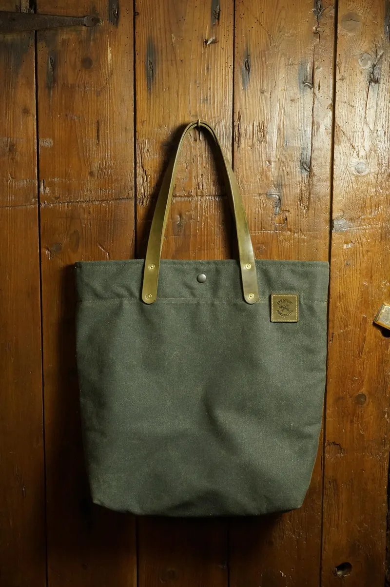 Khaki tote bag against a wooden wall, leather handles and leather patch with logo.