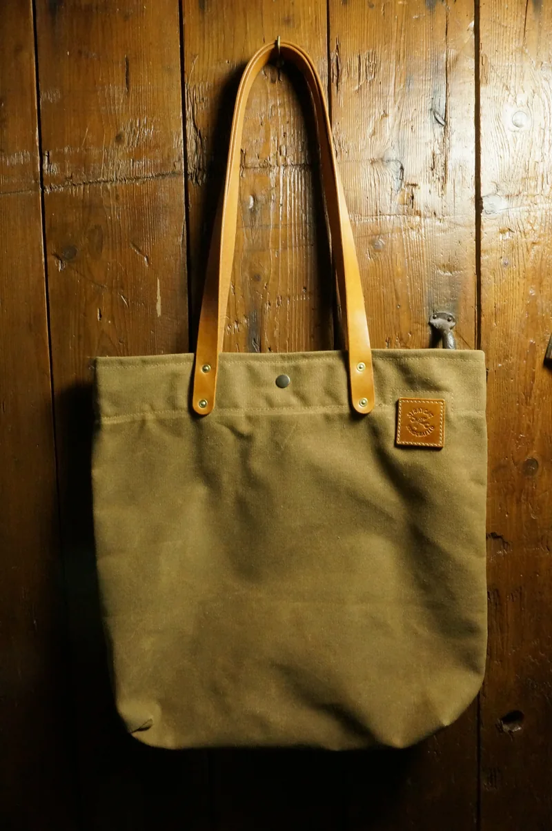 Ochre tote bag with light leather handles and leather patch, hanging against a wooden wall.