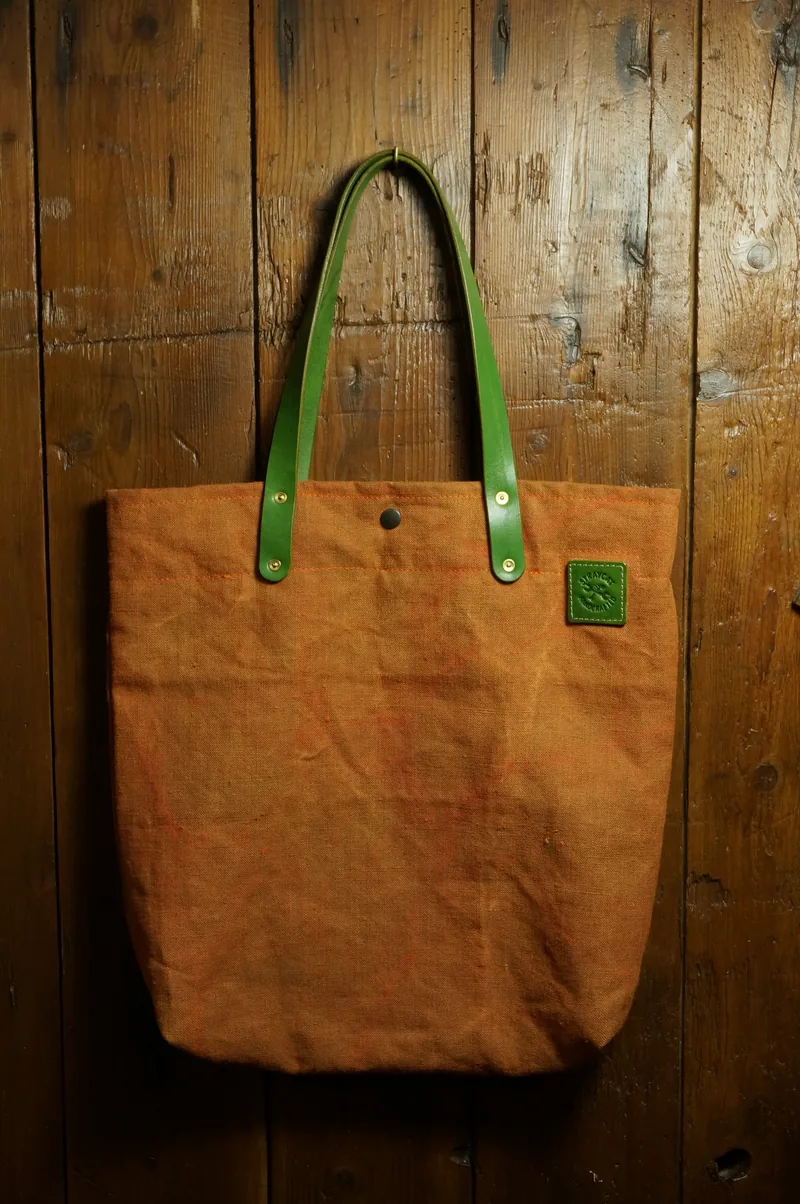Tent Orange tote bag with green handles and leather patch, hanging against a wooden wall.