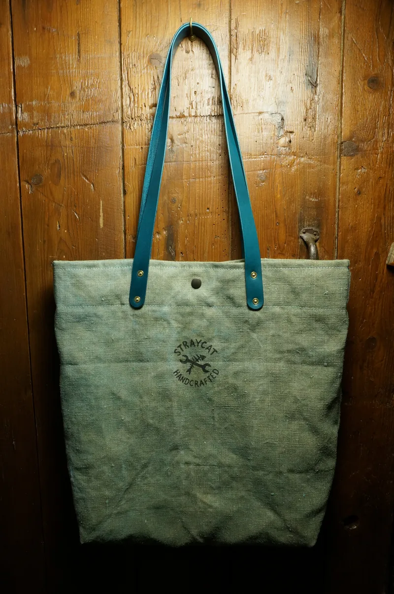 Tent Blue tote bag with turquoise handles and screen-printed logo, against a wooden wall.