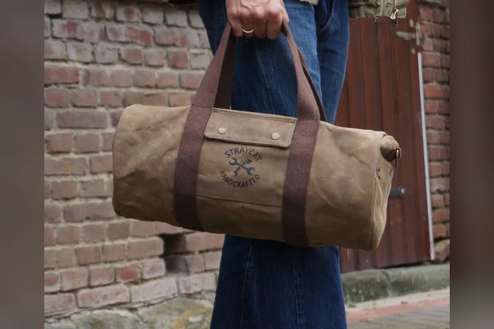 Duffle bag made from waxed canvas with screen-printed logo, carried by hand in front of a brick wall.